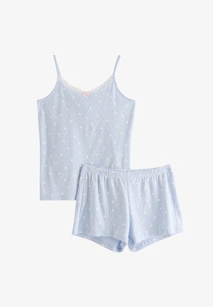 Ung kvinde med krøllet hår iført blå og hvide stribede ærmeløse top og matchende shorts står barfodet på en ensfarvet baggrund.