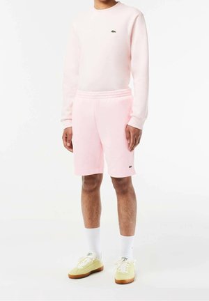 Person trägt hellrosa Sweatshirt und Shorts mit kleinen grünen Krokodil-Logos, weiße Socken und blassgelbe Sneakers, stehend auf weißem Hintergrund.