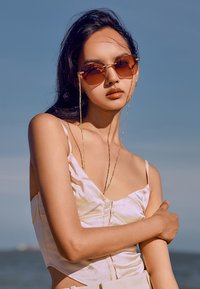 Occhiali da sole con lenti marroni rettangolari, dettagli in metallo dorato e una catena. Il modello indossa un top di raso rosa chiaro con spalline sottili.