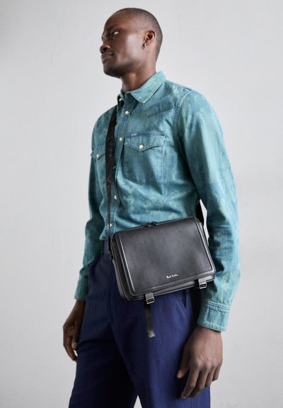 Borsa a tracolla in pelle nera con chiusura a zip, cinturino regolabile e due dettagli con fibbia, indossata sopra una camicia in denim verde e pantaloni blu navy.