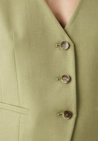 Grön väst med v-ringning, prydd med tre bruna knappar med tråddetaljer och en texturerad tygfinish. Inkluderar sidfickor.