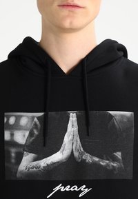 Black hoodie featuring a large monochrome print of hands in a prayer position, with intricate tattoos on the arms and white text "pray" at the bottom.
