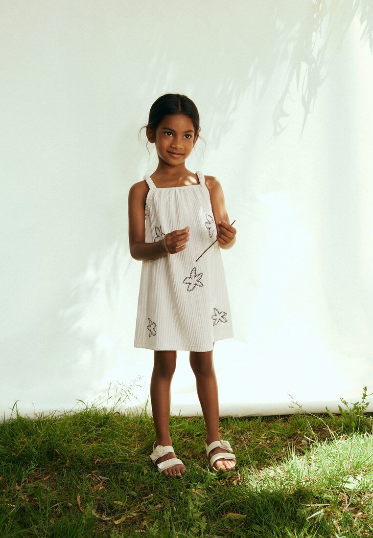 Fille en robe blanche sans manches avec des motifs floraux noirs, debout pieds nus sur l'herbe, tenant un bâton fin, souriant doucement sur un fond clair.