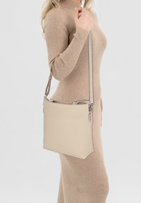 Woman in a beige ribbed turtleneck dress carrying a beige rectangular shoulder bag with an adjustable strap on her left side.