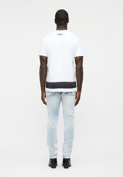 Άνδρας που φοράει λευκό κοντομάνικο μπλουζάκι με μαύρη και κόκκινη ρίγα κοντά στο τελείωμα, ανοιχτόχρωμο μπλε τζιν και μαύρα παπούτσια, όρθιος με την πλάτη στραμμένη.