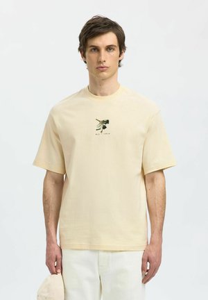 Beige bomulds T-shirt med korte ærmer, prydet med et grafisk print i midten. Modellen holder en matchende kasket i hånden, iført hvide bukser.