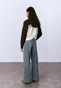 Suéter de punto trenzado en marrón con un panel blanco y beige; combinado con jeans grises holgados. El fondo es una superficie gris clara.