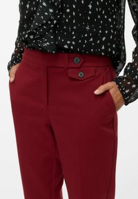 Mujer que lleva puestos unos pantalones burgundy ajustados con dos botones decorativos negros en la cintura y una blusa negra con un patrón y volantes.
