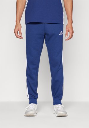 Mężczyzna w niebieskich spodniach dresowych Adidas z białymi paskami i białych sportowych butach, stojący na jednolitym jasnym tle.