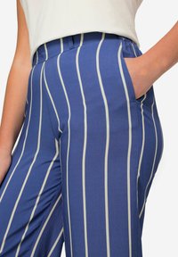 Modré pruhované kalhoty s upraveným střihem, které mají svislé krémové pruhy, boční kapsy a hladkou texturu látky.