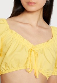 Blusa cropped amarela feita de um tecido leve. Apresenta um decote quadrado, mangas franzidas e um detalhe de laço na parte da frente.