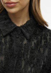 Melnā ar garām piedurknēm krekls, kas izgatavots no caurspīdīga auduma ar teksturētu, pūkainu virsmu, izteikta apkakle un priekšējie pogu elementi.