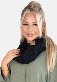 Étole noire texturée enroulée autour du cou, avec un bord effiloché. Portée avec une chemise verte à boutons et des accessoires dorés.