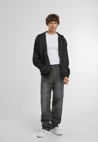 Schwarze Zip-Hoodie mit Kordeln, weißes T-Shirt darunter, graue weite Cargo-Hose mit Nähdetails und weiße Turnschuhe.