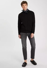 Czarny sweter na zamek z długimi rękawami, w zestawieniu z szarymi dżinsami. Model nosi granatową bandanę w wzory oraz czarne buty wsuwane.