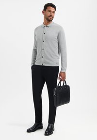 Grijze lange mouwen overhemd met knoopsluiting en kraag, zwarte knopen, gecombineerd met zwarte tailor-made broek en gepolijste zwarte laarzen, terwijl hij een zwarte aktentas vasthoudt.