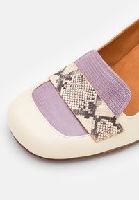 Mocassin crème avec un insert texturé lilas et une lanière en motif serpent traversant le orteil, montrant une partie de la semelle intérieure marron.