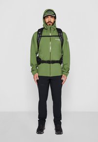 Chaqueta impermeable verde con capucha, correas negras y mochila, combinada con pantalones negros y botas de senderismo, adecuada para actividades al aire libre.