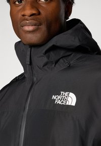 Crna kapuljača jakna izrađena od laganog materijala. Ima prednji zip zatvarač, podesivu kapuljaču i bijeli logo "The North Face" na prsima.