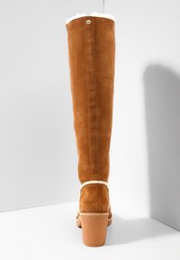 Botte haute en suède marron avec talon bloc et doublure en laine blanche visible en haut et autour de la cheville, photographiée de dos sur fond blanc.