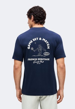 Mand iført navyblå t-shirt med hvidt tennis-tema grafik og tekst, der lyder ""Game Set & Match, French Heritage Lacoste Club"" på bagsiden.