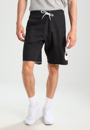 Mand iført sorte shorts med snøre og hvidt logo på siden, hvide sokker, hvide sneakers og en grå t-shirt, stående på en ensfarvet baggrund.