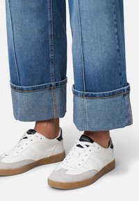 Białe sneakersy z teksturowanymi akcentami i gumowymi podeszwami, w połączeniu z szerokimi dżinsami, które mają surowe wykończenie i jasnoniebieski odcień.