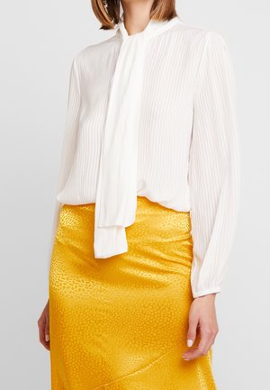 Kvinde iført en hvid bluse med lange ærmer og slips samt et skinnende gult mønstret skørt foran en ensfarvet baggrund.