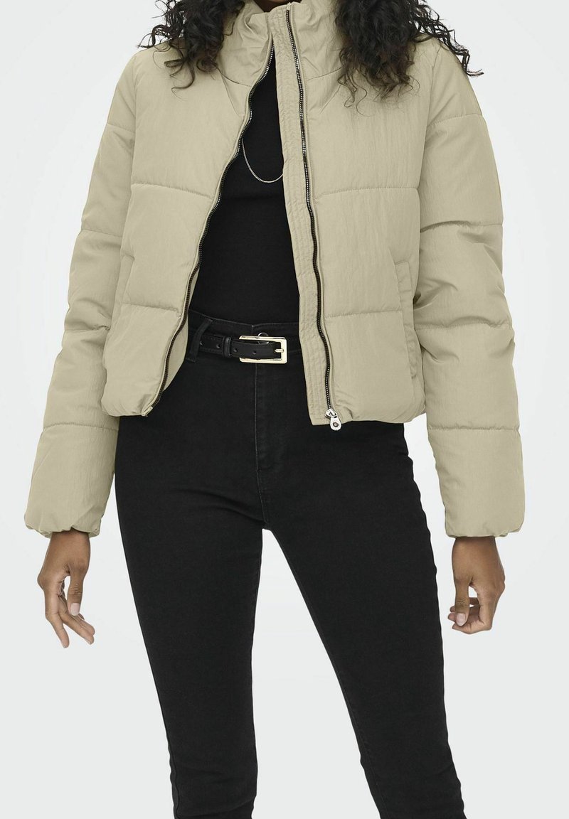 Femme portant une veste matelassée beige sur un haut noir et un pantalon noir taille haute avec une ceinture noire, debout devant un fond uni.