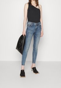 Top negro de un solo hombro, vaqueros ajustados de color azul claro con ligero desvanecimiento y mules negros de tacón. La modelo lleva un bolso de cuero negro.