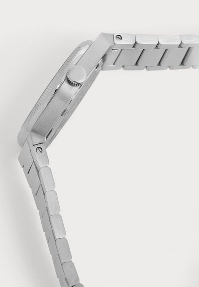 Vue latérale d’une montre-bracelet en métal argenté avec une couronne texturée et un bracelet à maillons sur un fond uni clair.