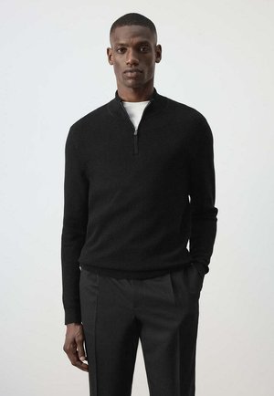 Uomo che indossa un maglione nero con cerniera e maniche lunghe e pantaloni neri, in piedi davanti a uno sfondo chiaro e uniforme.