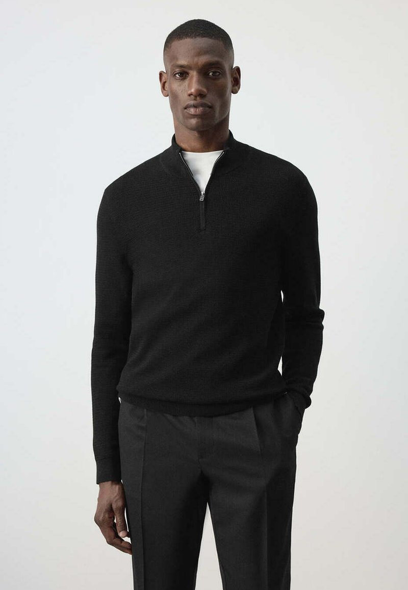 Homme portant un pull noir zippé à manches longues et un pantalon noir, debout devant un fond clair uni.