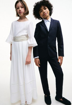 Ragazza giovane in un lungo vestito bianco con pizzo e ragazzo in un abito scuro con camicia bianca stanno fianco a fianco contro uno sfondo semplice.