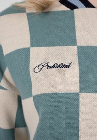 Nærbillede af en grøn- og beige ternet sweater med ordet "Forbudt" broderet i kursiv på brystet.