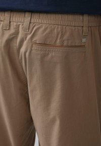 Braune Baumwollhosen mit elastischem Bund, ausgestattet mit einer einzelnen Gesäßtasche und einem kleinen metallischen Logo-Detail. Glatte Textur, lässiges Design.