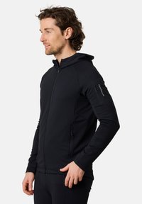 Felpa nera con zip e cappuccio, realizzata in un tessuto testurizzato, con silhouette aderente, tasche laterali e una piccola tasca con zip sul braccio sinistro.