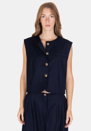 Donna con lunghi capelli castano chiaro indossa un gilet senza maniche blu navy con grandi bottoni e pantaloni a vita alta abbinati su sfondo bianco.
