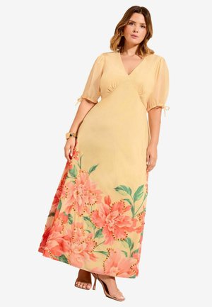 Woman wearing a yellow wrap dress with sheer puff sleeves and large pink floral print at the hem, paired with beige high-heeled sandals.