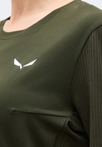 Κοντινό σε πουκάμισο ελιάς με λευκό γωνιακό λογότυπο στο στήθος και υφασμάτινη υφή στο μανίκι.