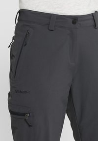 Pantalones de exterior para hombre en gris oscuro, fabricados en material suave. Presentan múltiples bolsillos con cremallera y un logo en el muslo izquierdo. Cintura ajustable.
