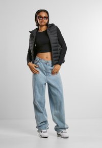 Crna puffer prsluk preko crnog crop topa, uparen sa svijetloplavim širokim trapericama i bijelim tenisicama. Model nosi tamne sunčane naočale.