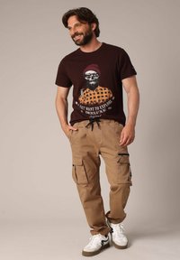 Camiseta gráfica marrón con un diseño de calavera y cuadros. Pantalones cargo beiges con múltiples bolsillos y cremalleras. Zapatillas blancas con acentos negros.