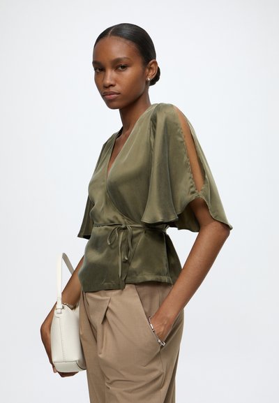 Femme portant un chemisier cache-cœur vert olive à manches ouvertes et un pantalon beige taille haute, tenant un sac à main blanc.