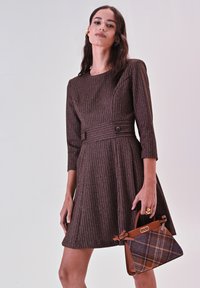 Bruine textuur jurk met driekwartmouwen, taille-riem en uitlopende rok, gecombineerd met een bruine handtas met ruitpatroon.