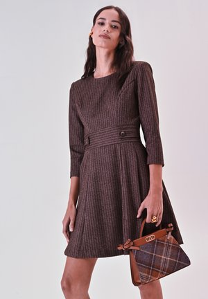 Bruine textuur jurk met driekwartmouwen, taille-riem en uitlopende rok, gecombineerd met een bruine handtas met ruitpatroon.