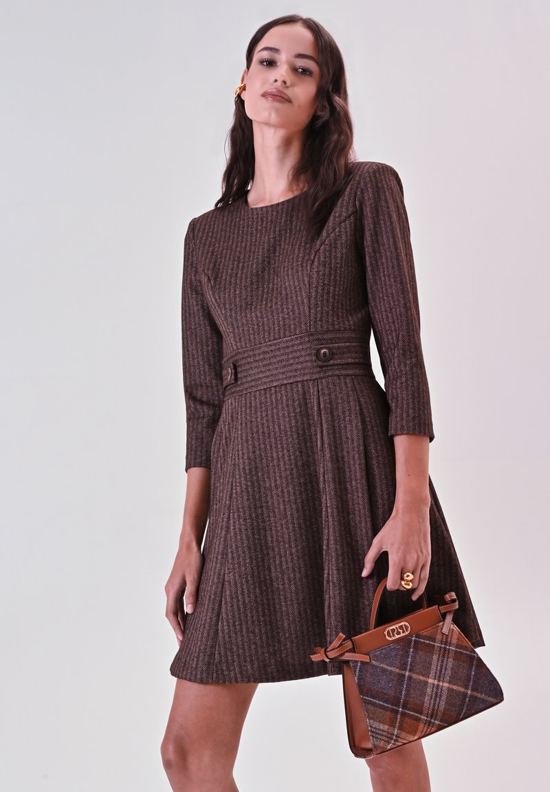 Bruine textuur jurk met driekwartmouwen, taille-riem en uitlopende rok, gecombineerd met een bruine handtas met ruitpatroon.