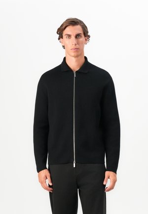 Young man with short wavy hair wears a black zip-up sweater and black pants, standing against a plain white background.
