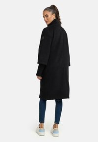 Zwarte, lange jas met een hoge kraag, relaxte pasvorm en opgerolde mouwen, gecombineerd met blauwe slim-fit jeans en witte sneakers. Heeft een logodetail op de mouw.