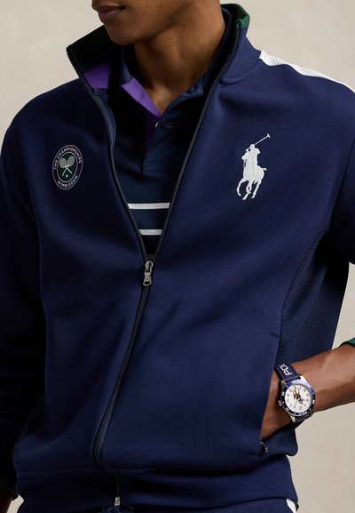 Tmavomodrá bunda na zips s bielymi detailmi a logom Wimbledonu, oblečená cez fialovo pruhované polo tričko. Dopĺňa ju strieborné hodinky s modrým remienkom.